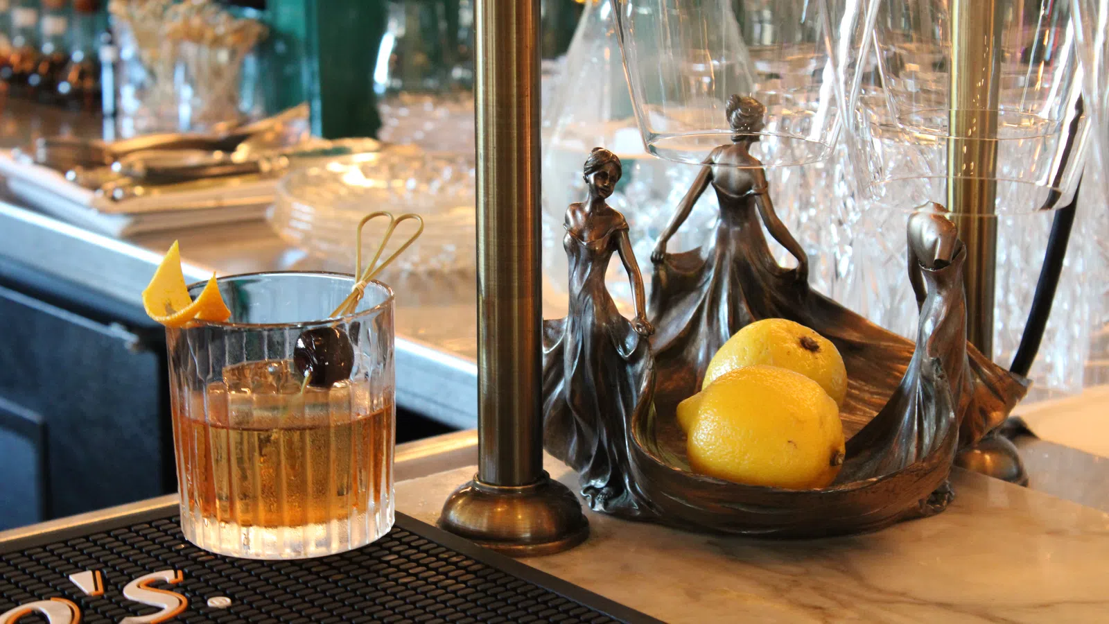 Whiskey cocktail with orange peel and cherry next to an art deco lemon holder shaped as dancing ladies on bar counter.