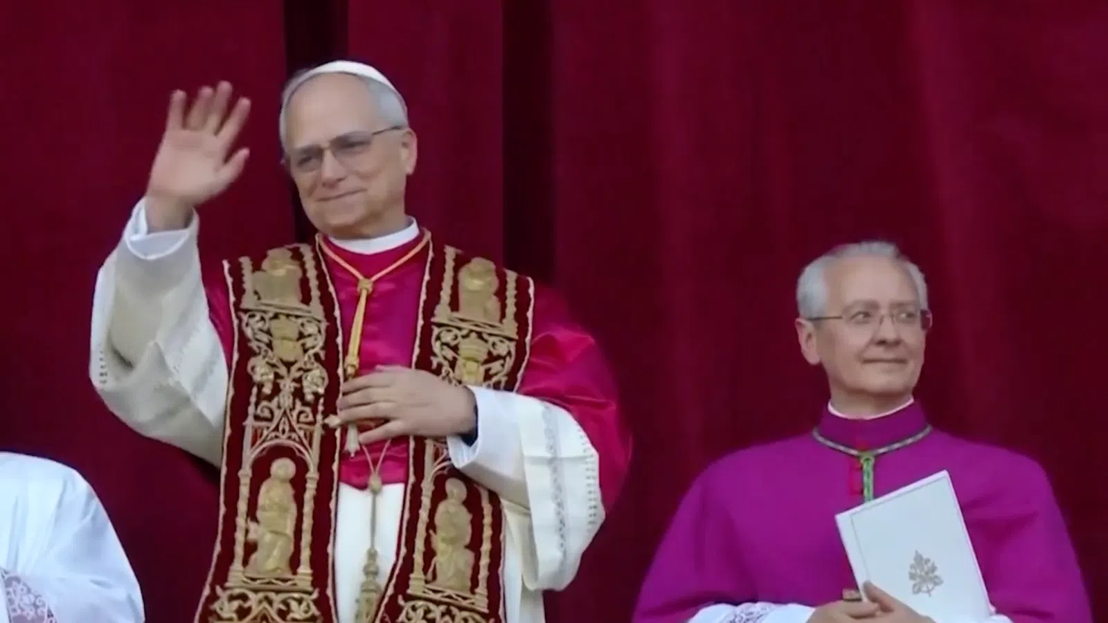 Pope in ornate vestments waving with two clergymen in ceremonial robes against red curtain background.