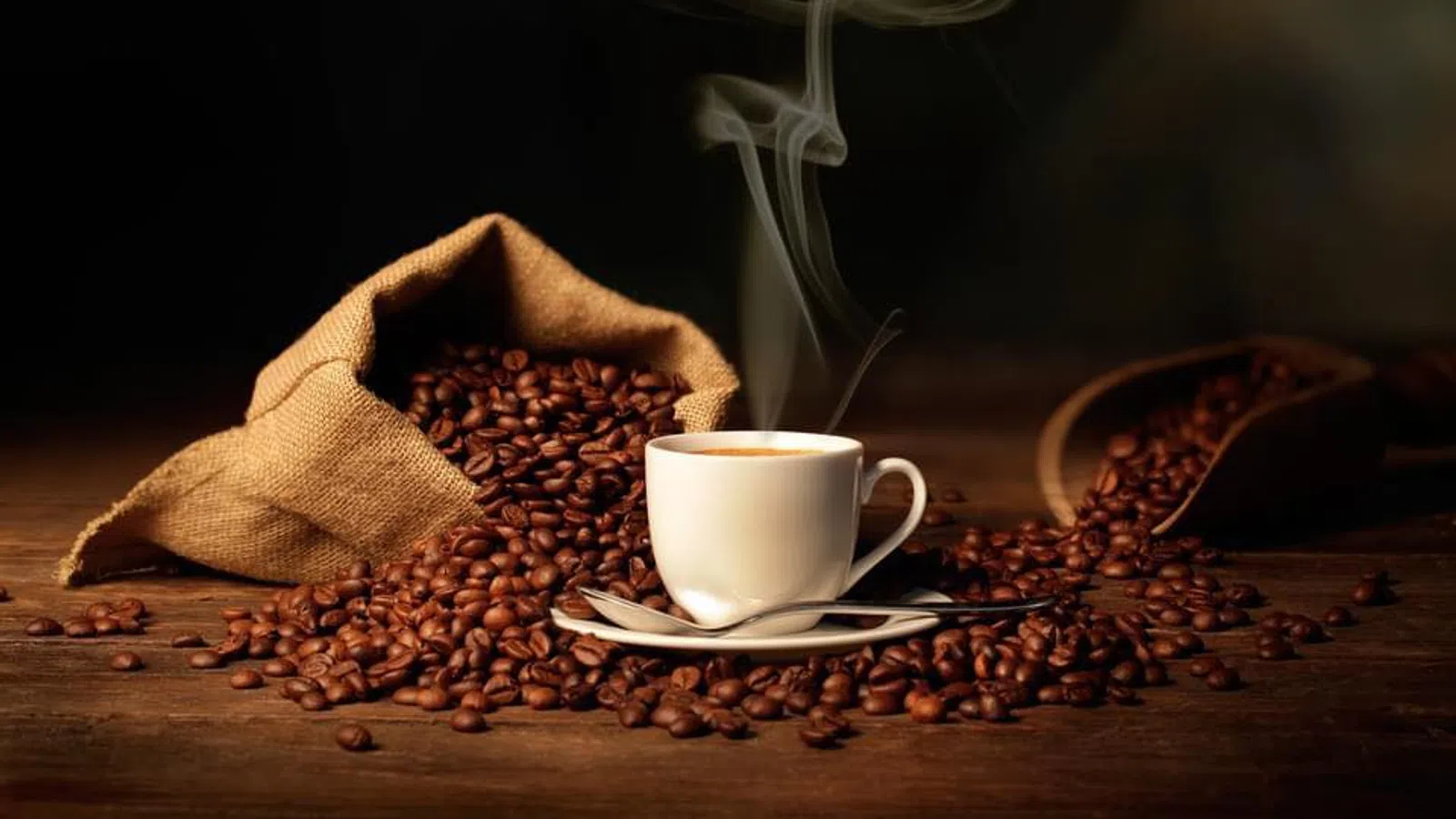 Steaming cup of coffee surrounded by spilled coffee beans from burlap sacks on a wooden surface.