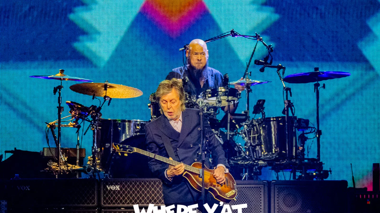 Paul McCartney playing bass guitar with drummer behind him on stage during a concert with a colorful background.
