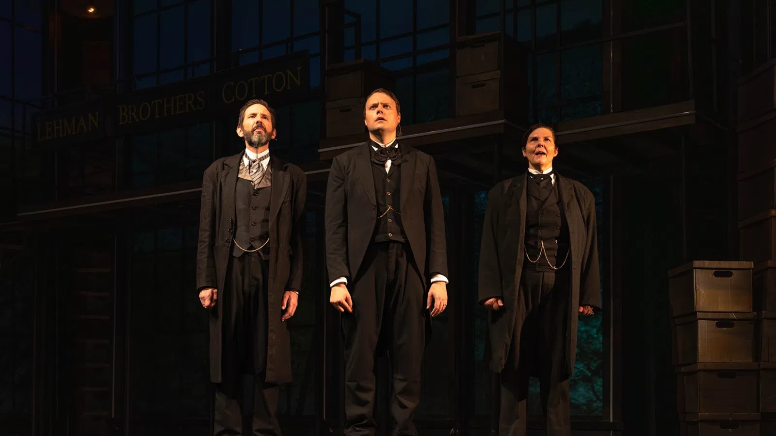 Three men in vintage suits stand on stage in front of an industrial backdrop under warm lighting during a theater performance.