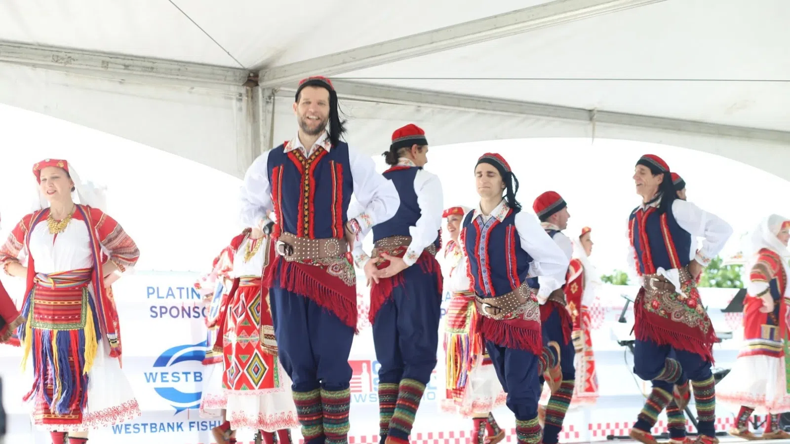 Group of dancers performing traditional folk dance in colorful Eastern European costumes under a white tent.