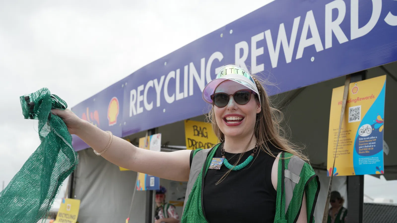 Woman in a green vest at a recycling event, promoting recycling rewards with enthusiasm.