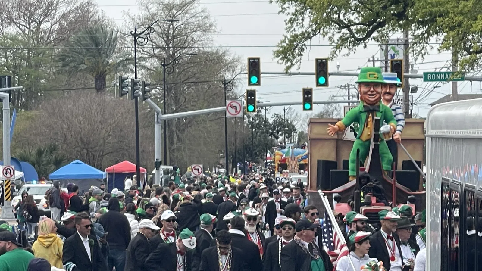Crowd in green attire and suits parading down city street with large leprechaun float during festive event.