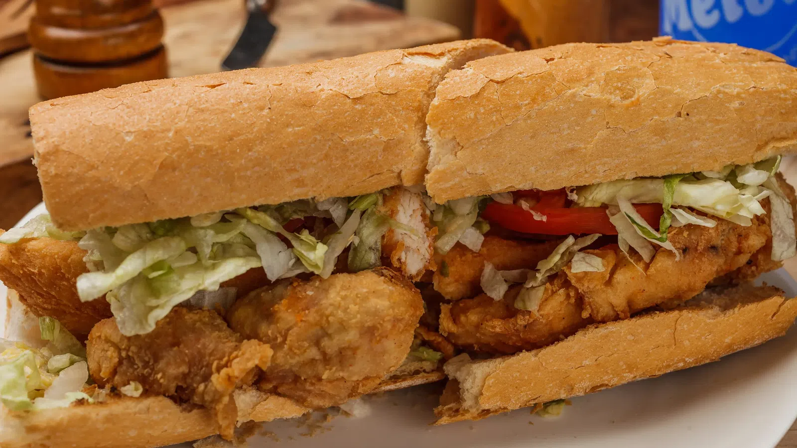 Crispy fried chicken po'boy sandwich with lettuce and tomato on a crusty French bread roll on a white plate.