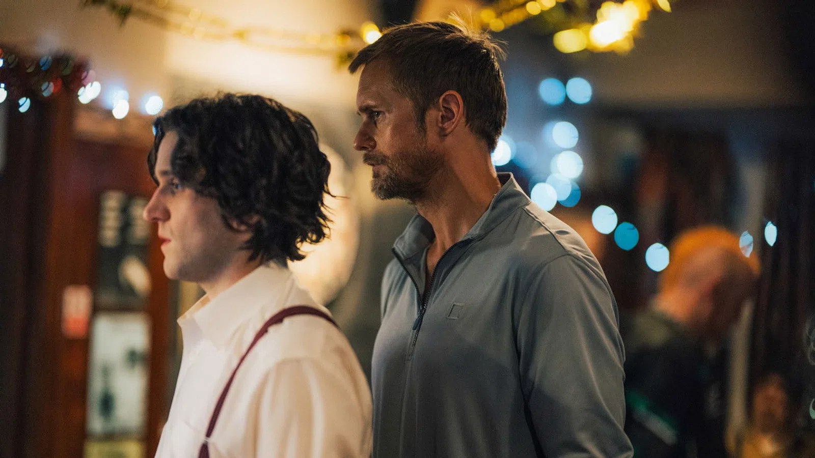 Two men standing side by side indoors with blurred lights and people in background