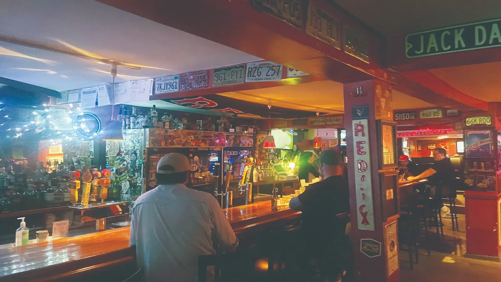 Interior of a cozy bar with patrons seated at the wooden counter, vintage signs, and warm lighting.