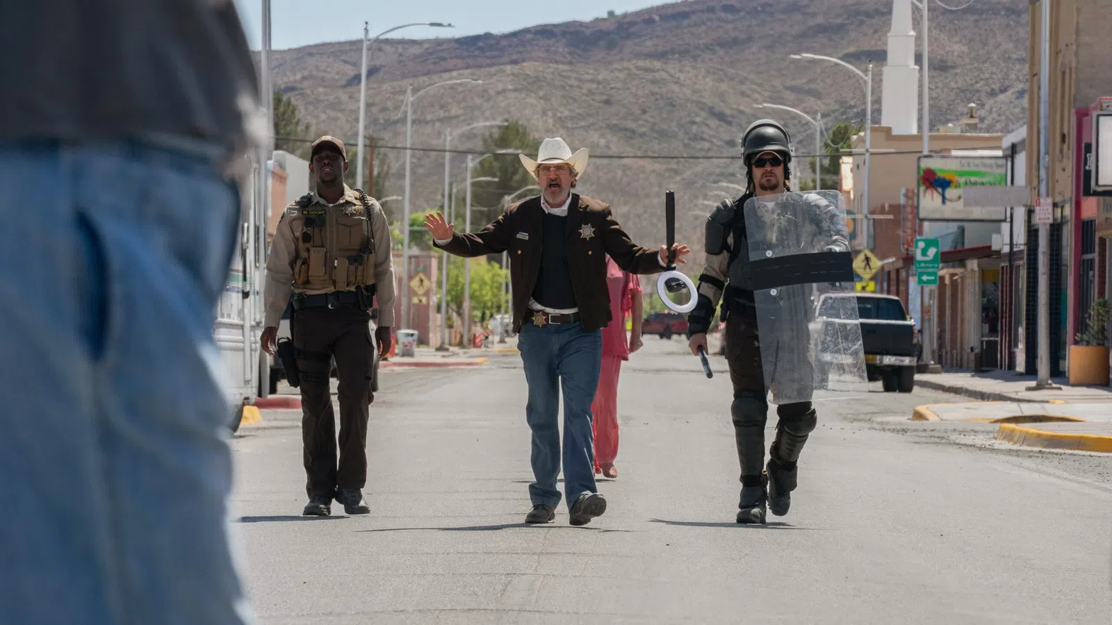 Sheriff and law enforcement officers walking down a sunny street in a desert town with mountains in background