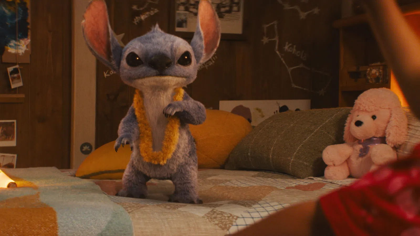 Blue alien creature wearing a yellow lei standing on a bed in a cozy room with photos and a stuffed dog toy.