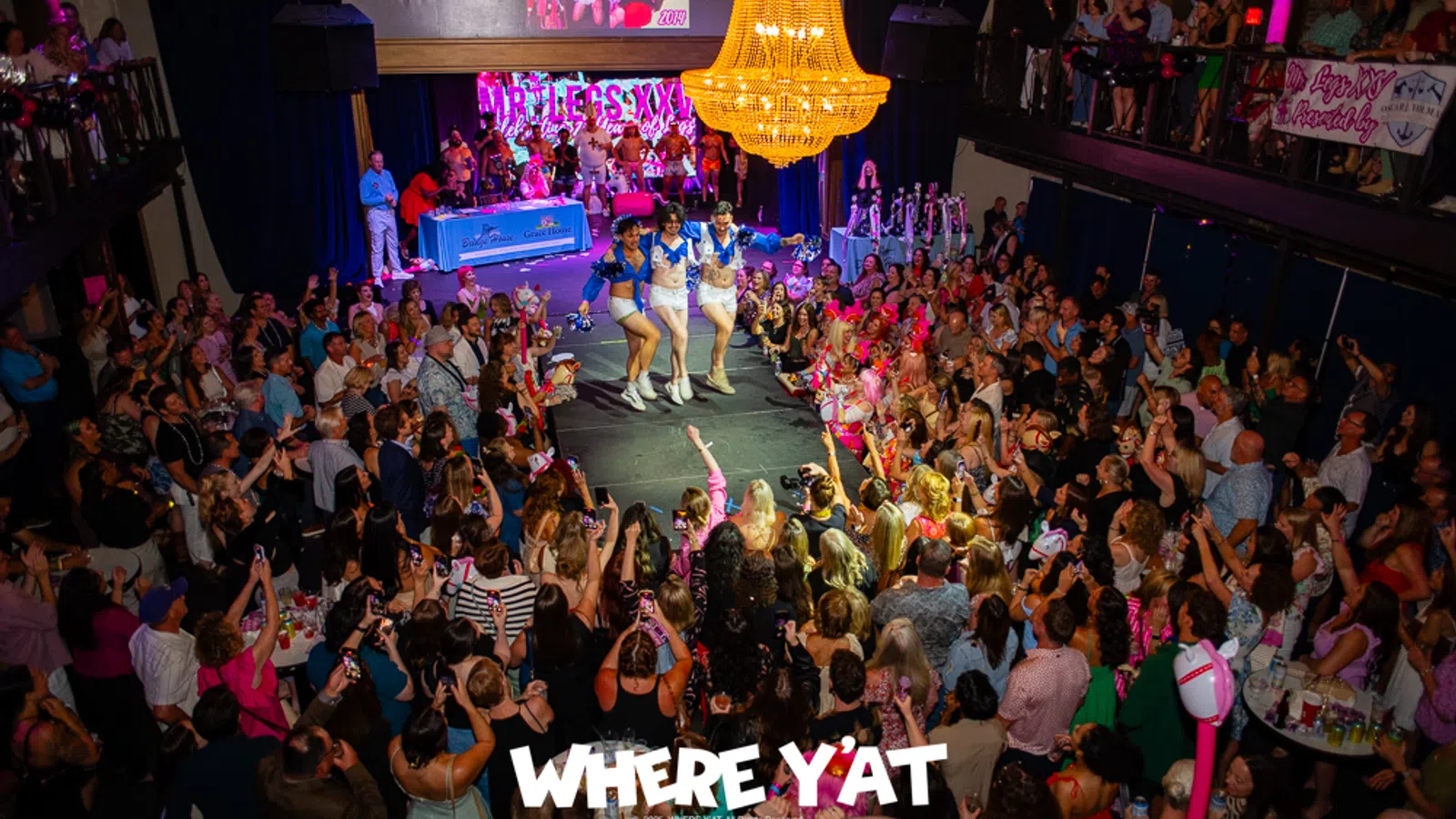 Crowded event with performers dancing on stage under a large chandelier in a vibrant venue packed with an enthusiastic audience.