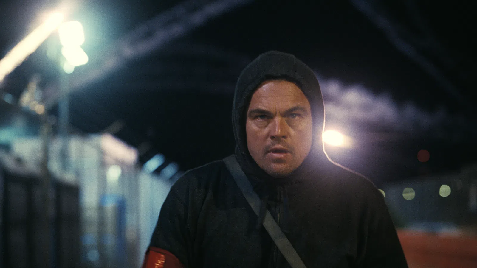 Man in dark hoodie with red armband walking at night in an urban area under bright streetlights.