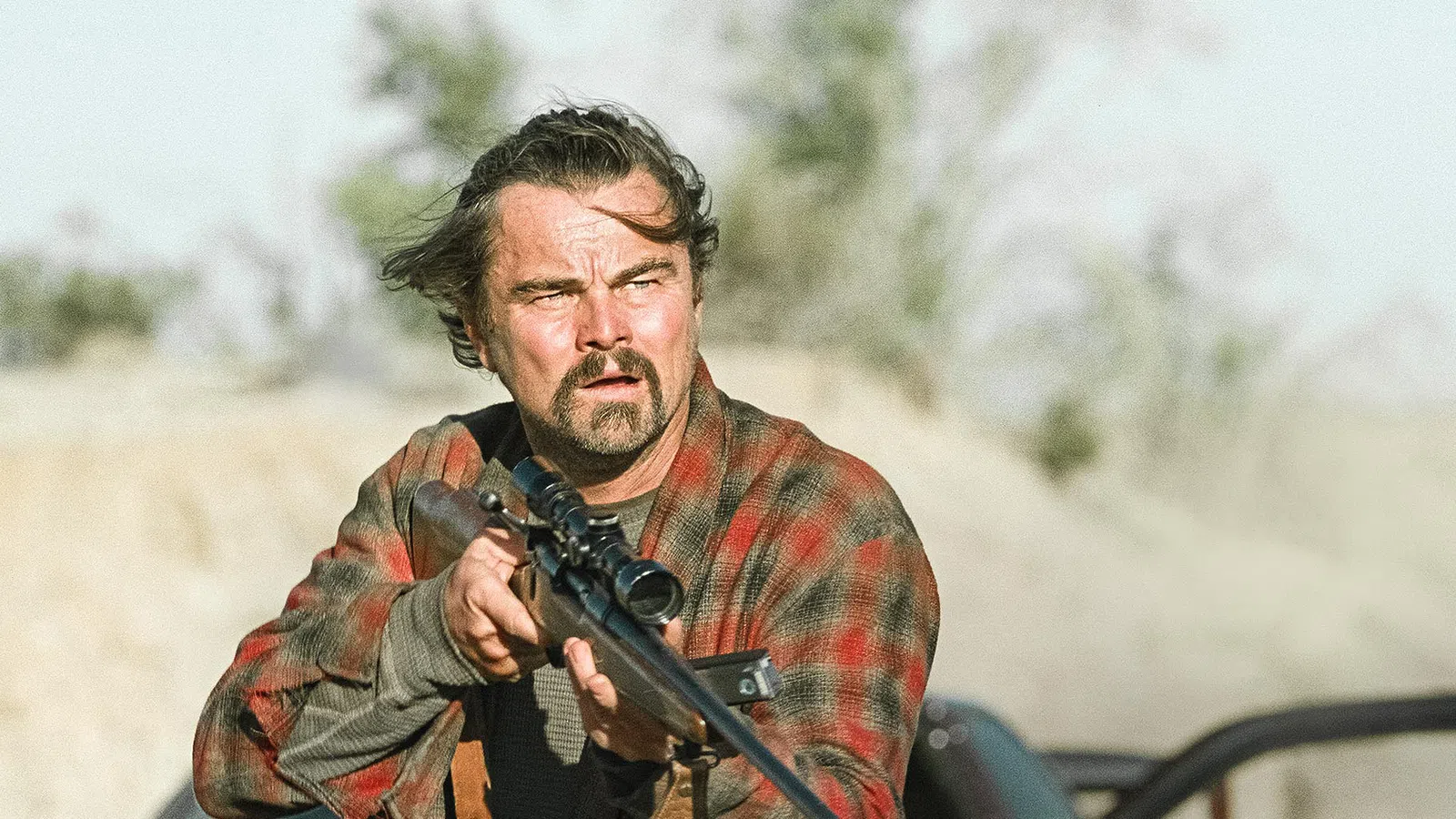 Man aiming a scoped hunting rifle outdoors wearing a red plaid shirt with intense focused expression.
