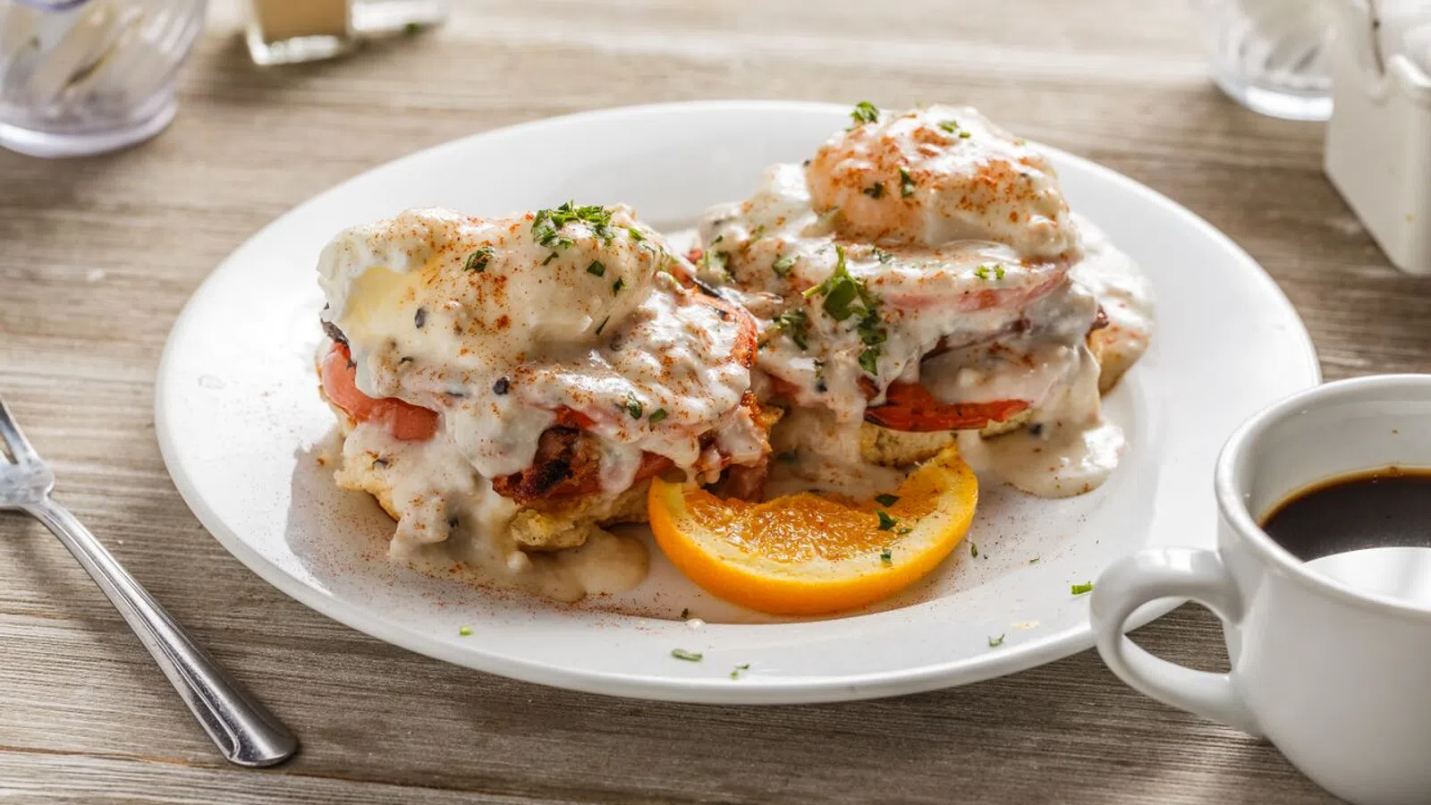 Plate of eggs Benedict with creamy sauce and herbs, garnished with an orange slice, next to a cup of black coffee.