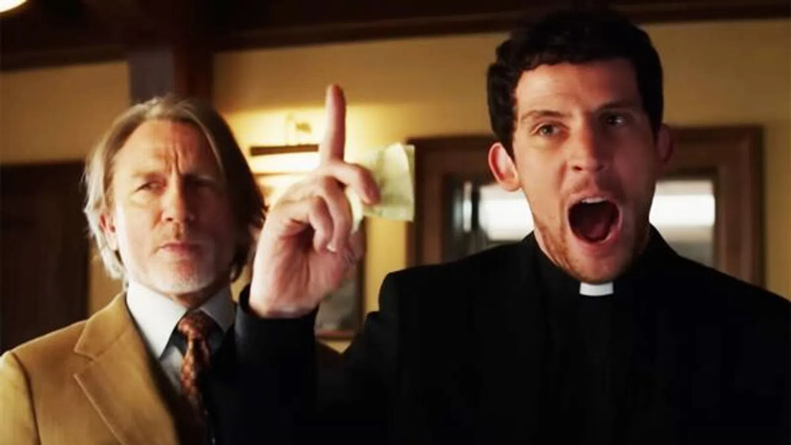 Man in black clerical collar raising finger and shouting while older man in tan suit watches in room.