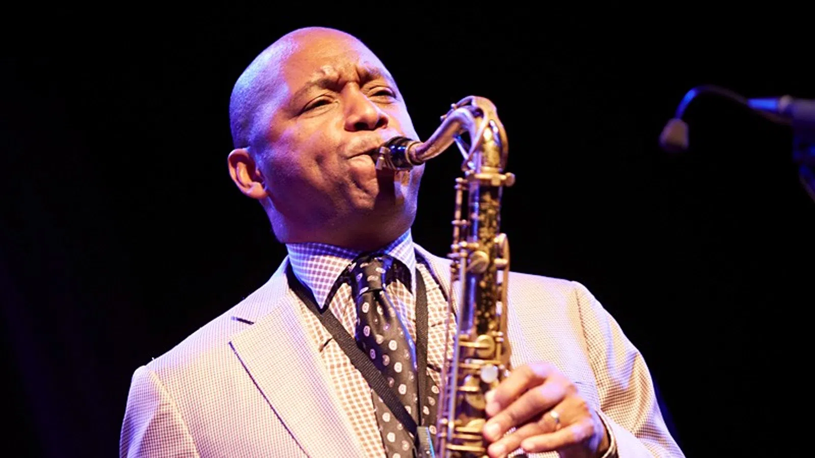 Jazz musician passionately playing saxophone on stage in a light-colored suit and patterned tie.