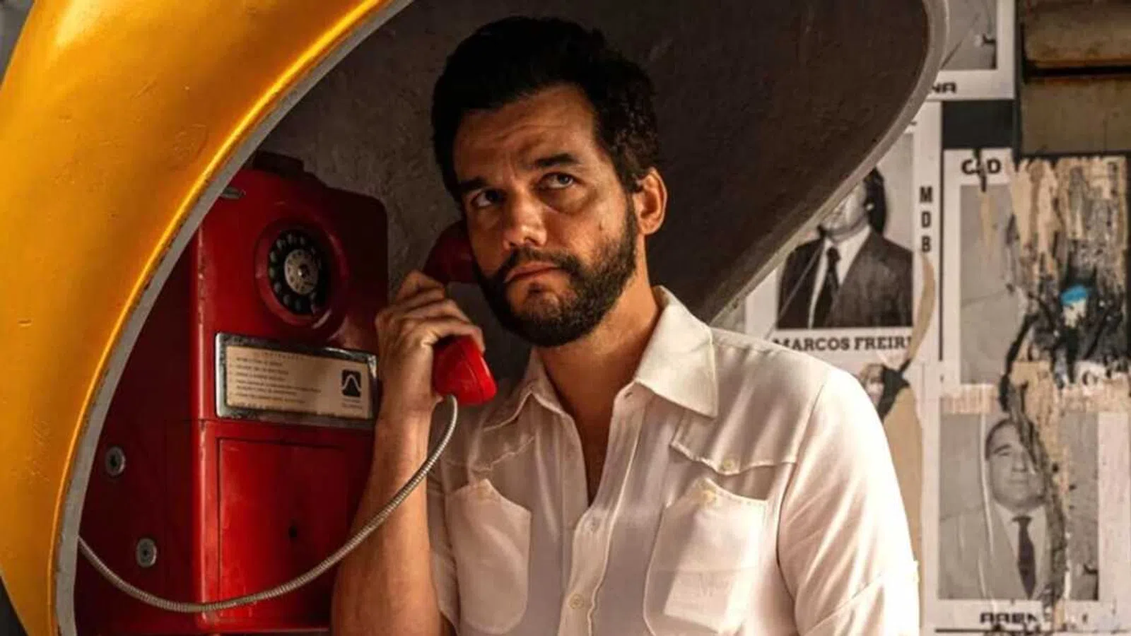 Man with beard using a red vintage phone booth with political posters on the wall behind him.