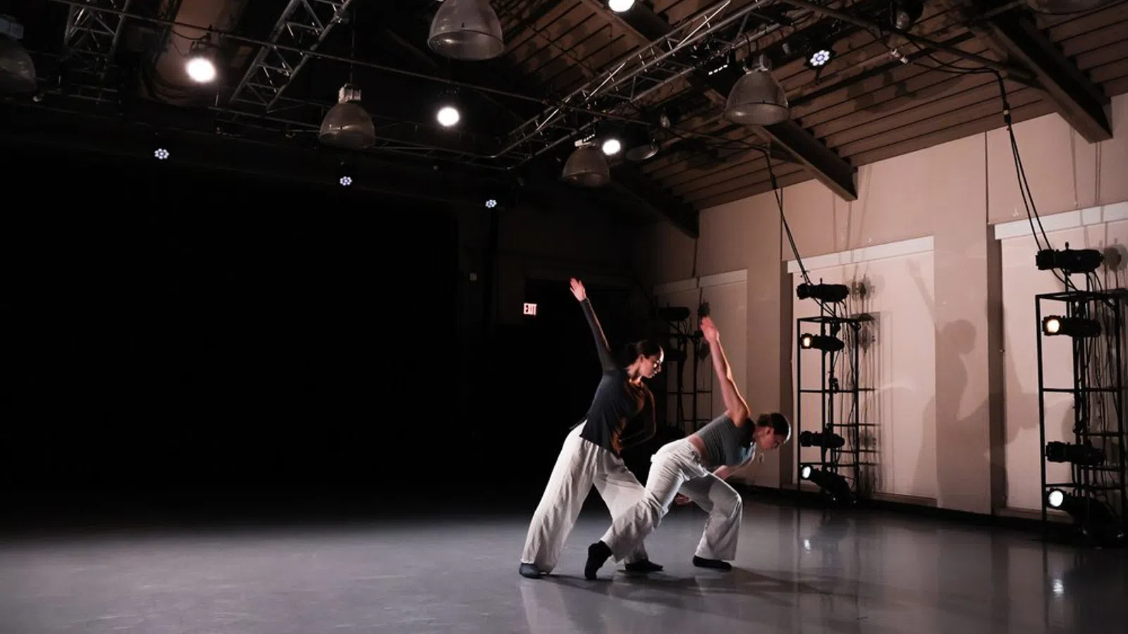 Two contemporary dancers performing in a dimly lit studio with spotlight and industrial ceiling beams.
