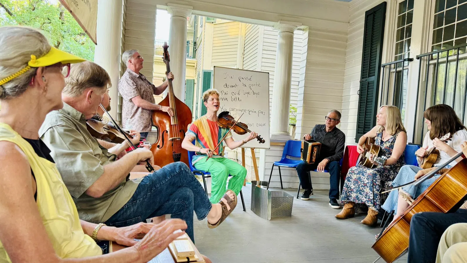 Group of people playing various musical instruments together on a covered porch during daylight.
