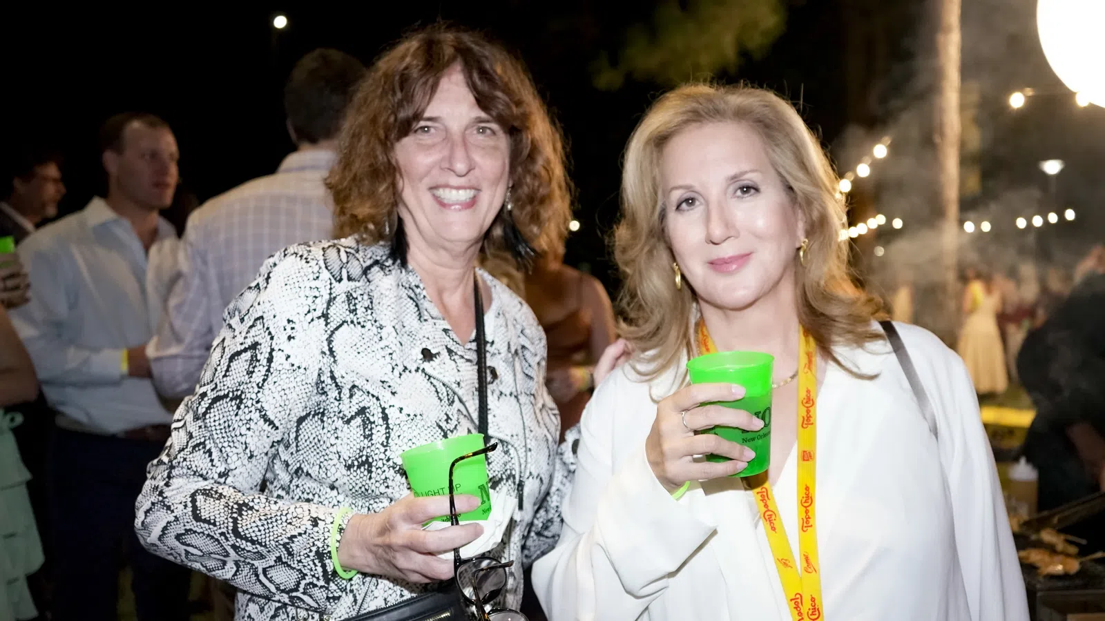 Two women smiling and holding green cups at an outdoor nighttime social event with string lights in background