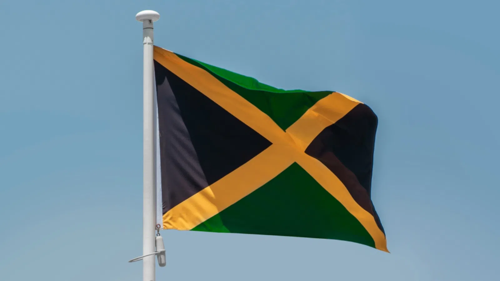 Jamaican flag waving on a white flagpole against a clear blue sky background.
