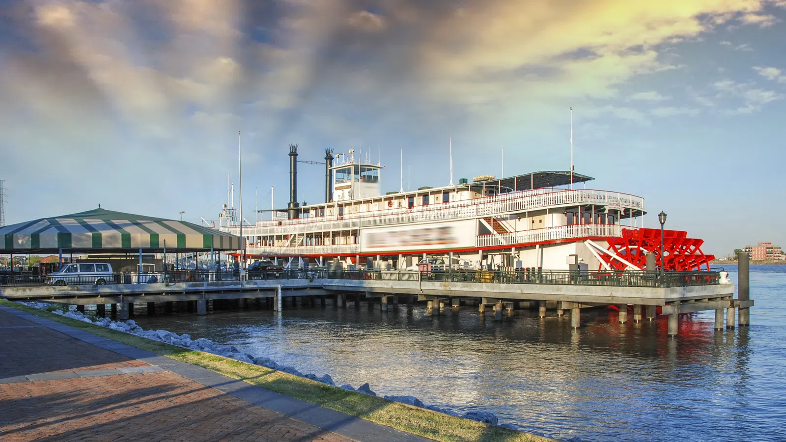 Historic riverboat docked at pier with red paddlewheel under a partly cloudy sky at sunset.