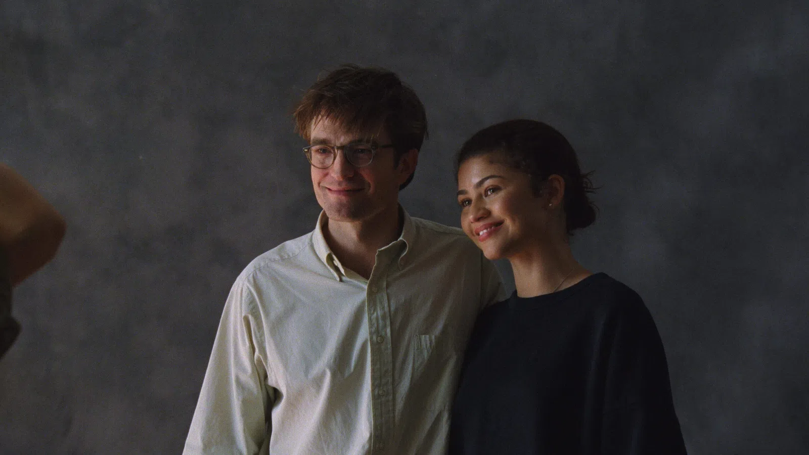 Smiling man with glasses and woman posing together against a dark gray textured background.