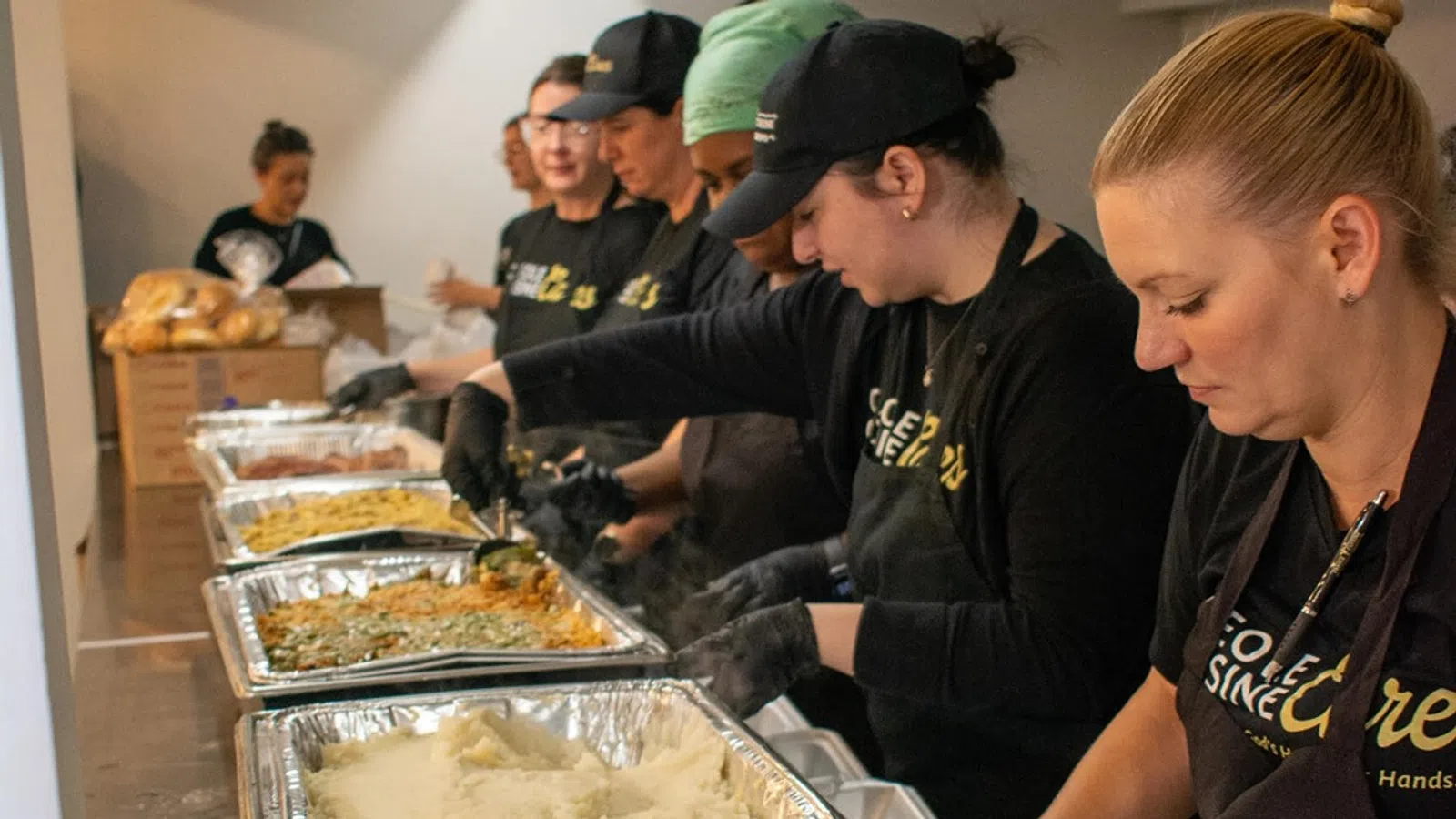 Volunteers wearing black gloves and aprons serve food from large trays in a community meal line.
