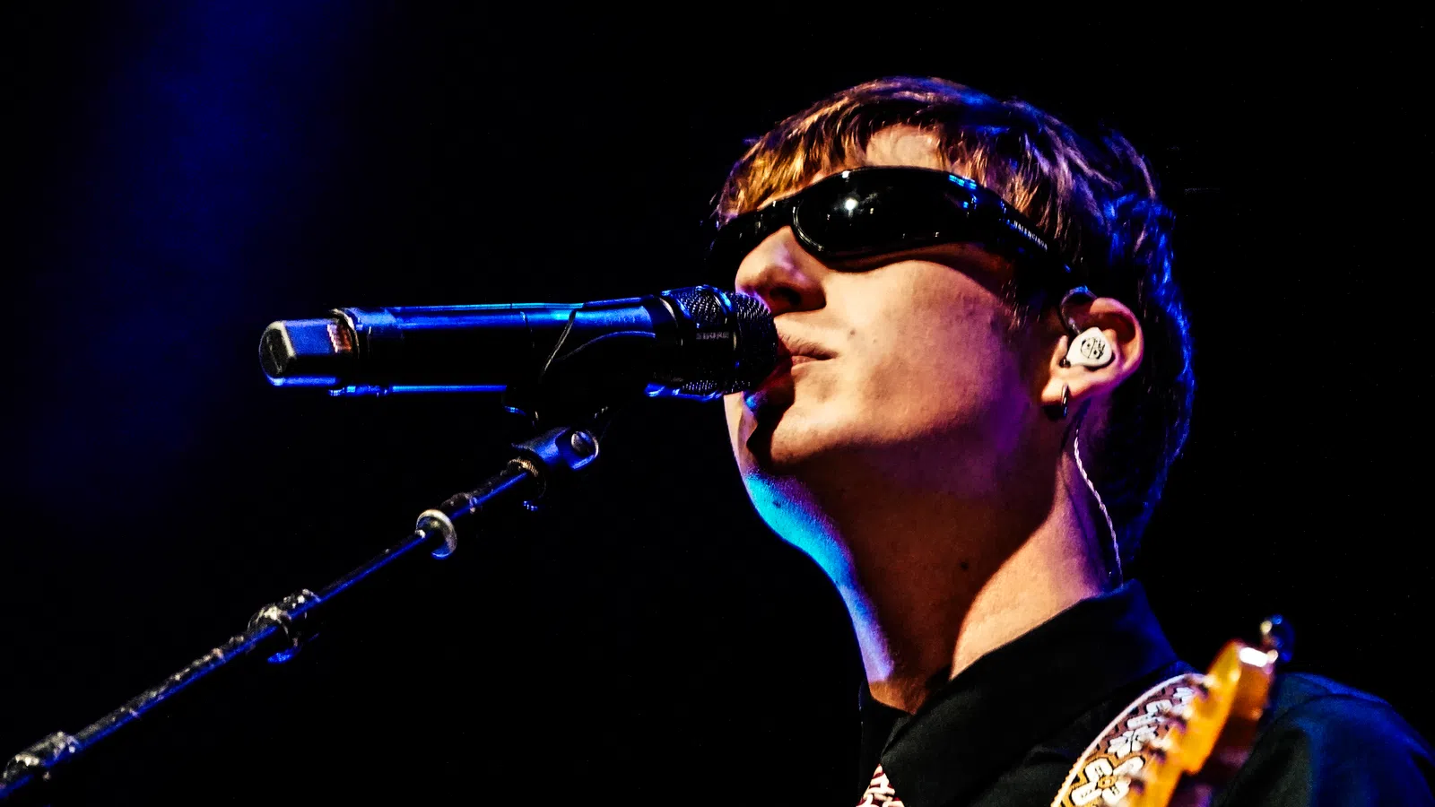 Male musician wearing sunglasses singing into microphone on stage with guitar under blue lighting
