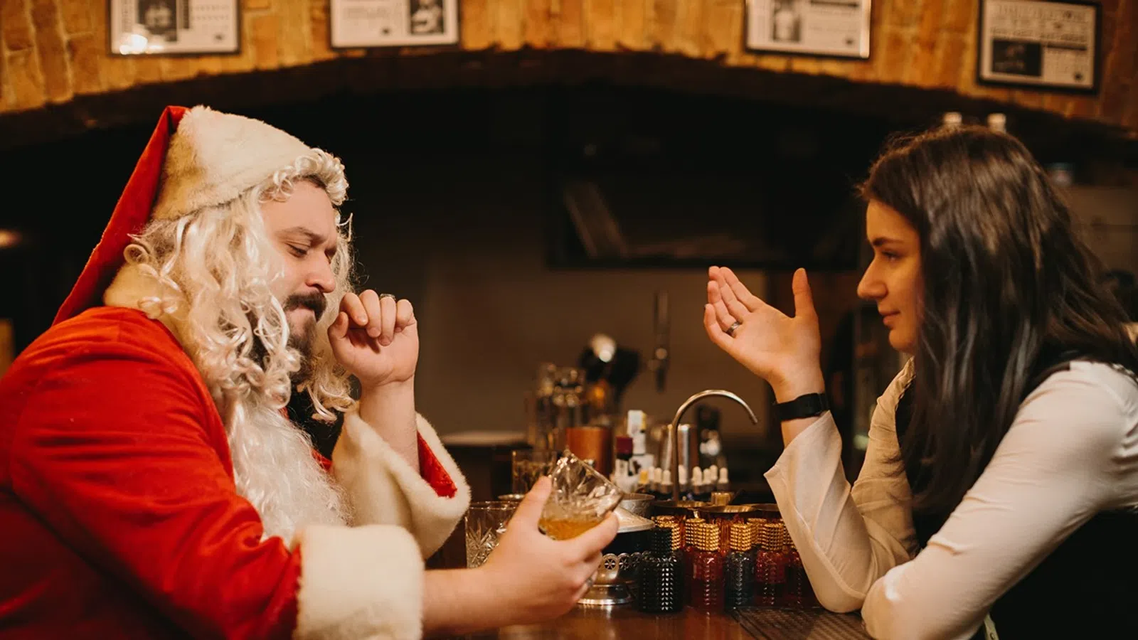 Man dressed as Santa drinking whiskey and talking to a woman at a dimly lit bar.