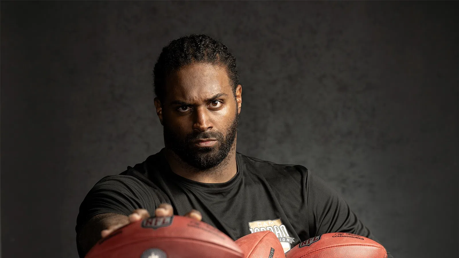 Serious male football player holding three footballs wearing a black shirt against a dark background