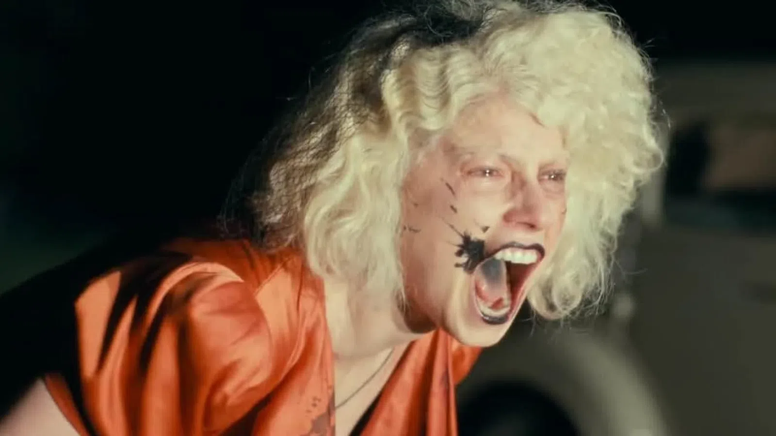 Person with wild blonde hair and black paint splatter on face yelling with mouth wide open in intense expression