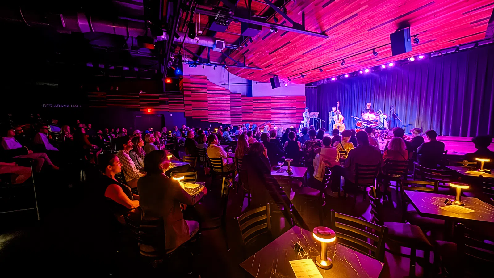 Audience seated in a dimly lit jazz club watching live band perform on stage with purple and pink lighting.