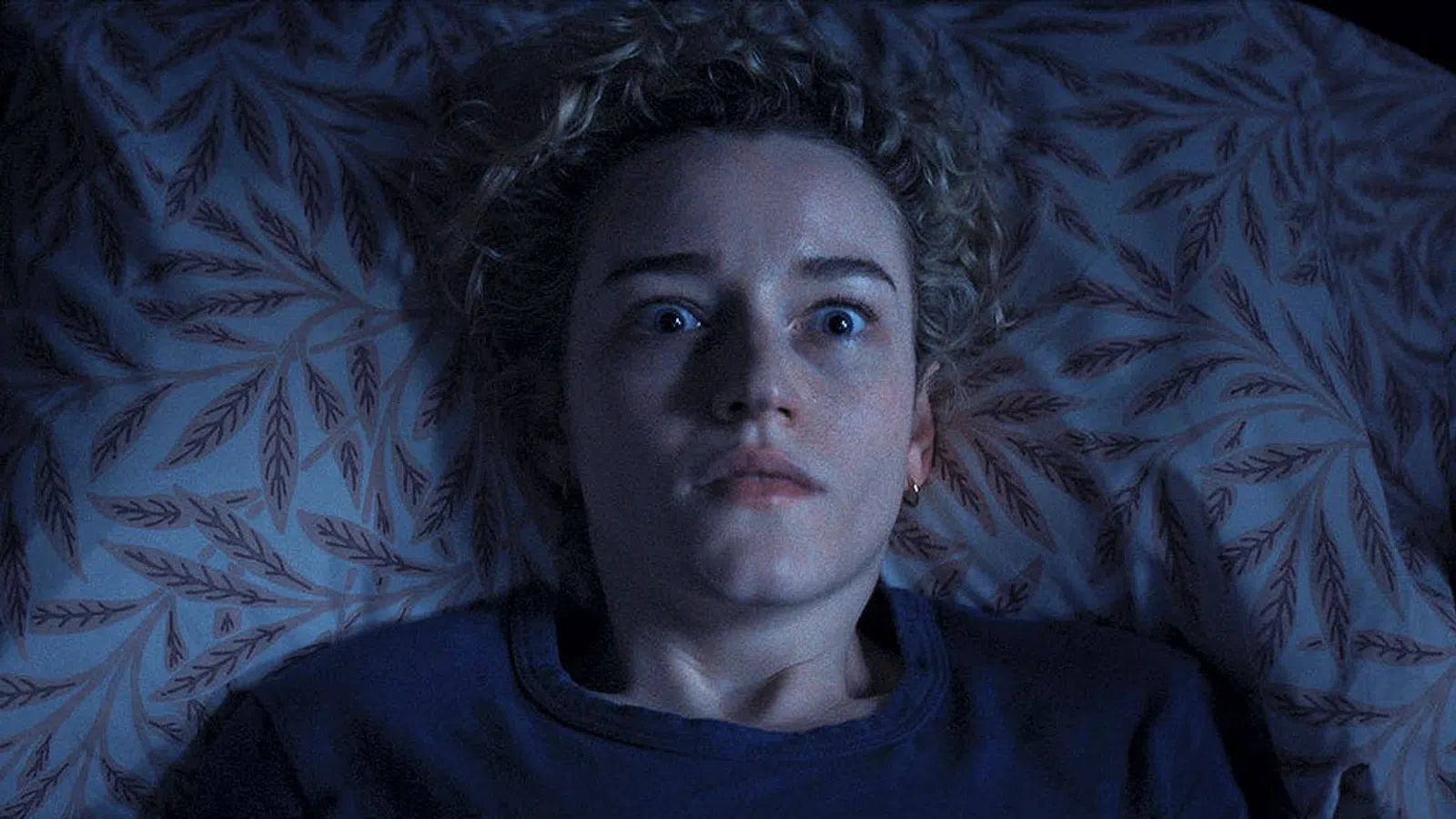 Woman with wide eyes lying in bed looking startled or frightened in dim blue lighting