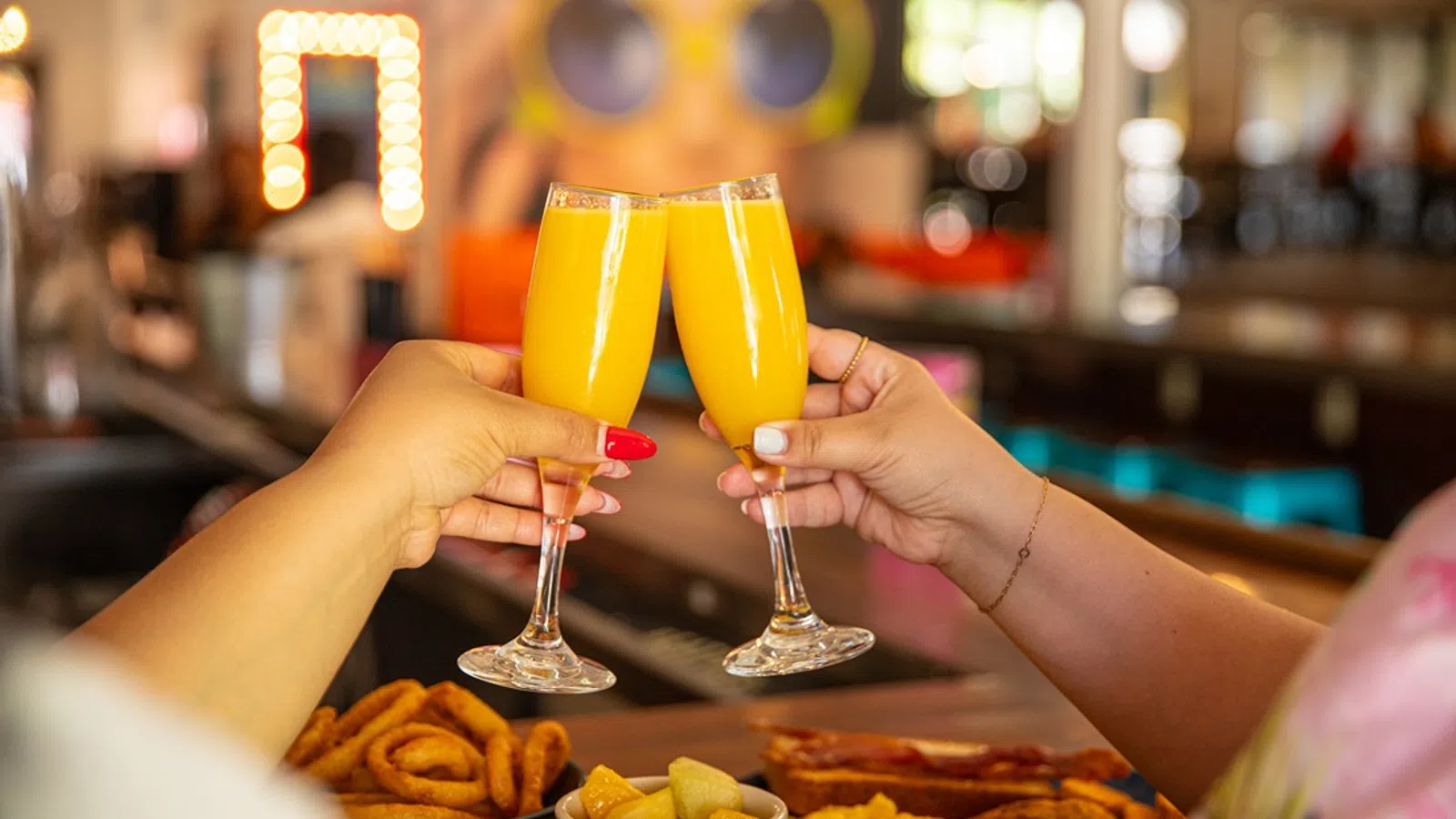 Two people clinking glasses of orange mimosa cocktails over a table with snacks in a cozy bar setting
