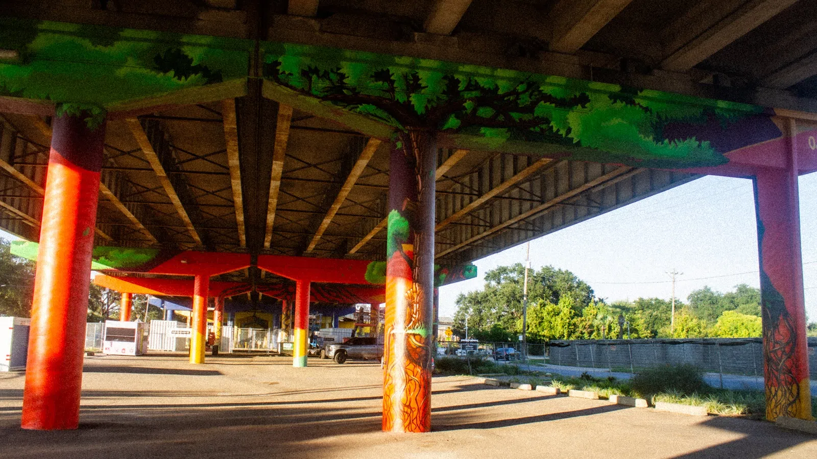Colorful mural of trees painted on the columns supporting a highway overpass in a sunny urban area.