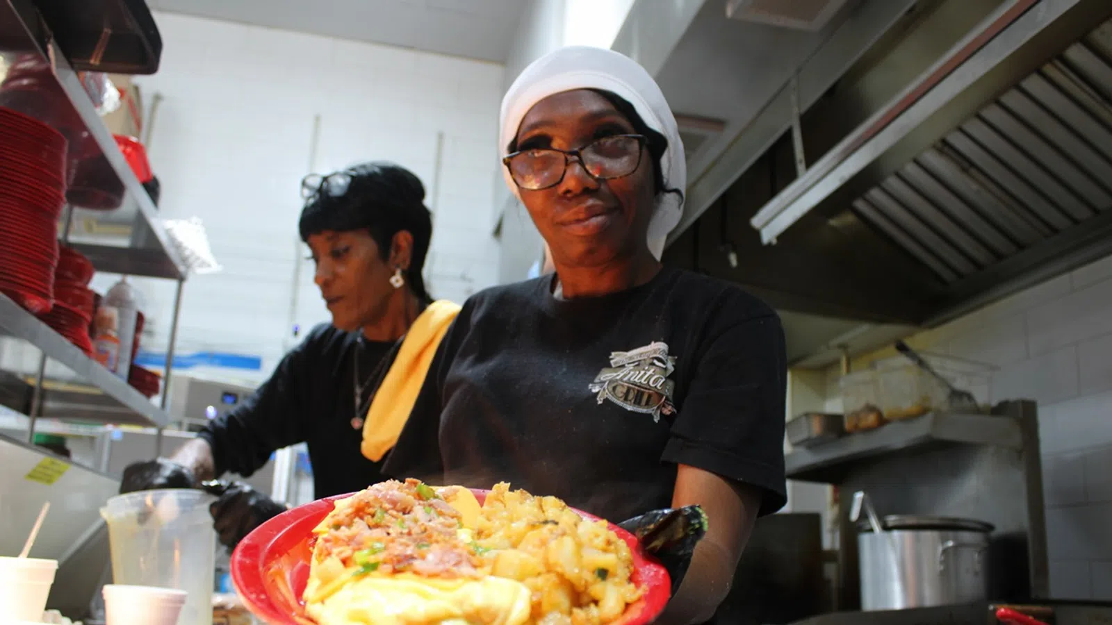 Smiling chef in kitchen holding a red plate with breakfast food including eggs, potatoes, and bacon.