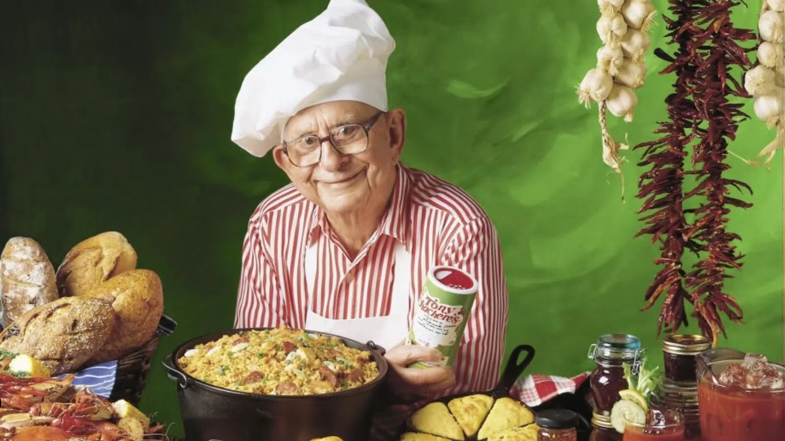 Elderly chef in white hat and red striped shirt presenting a large dish surrounded by bread, seafood, and vegetables.