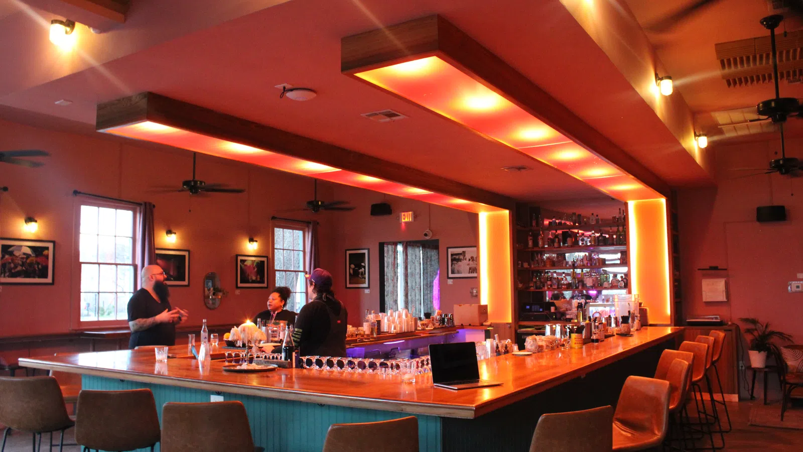 Modern bar interior with warm lighting, patrons chatting at L-shaped wooden counter, and high chairs surrounding it.