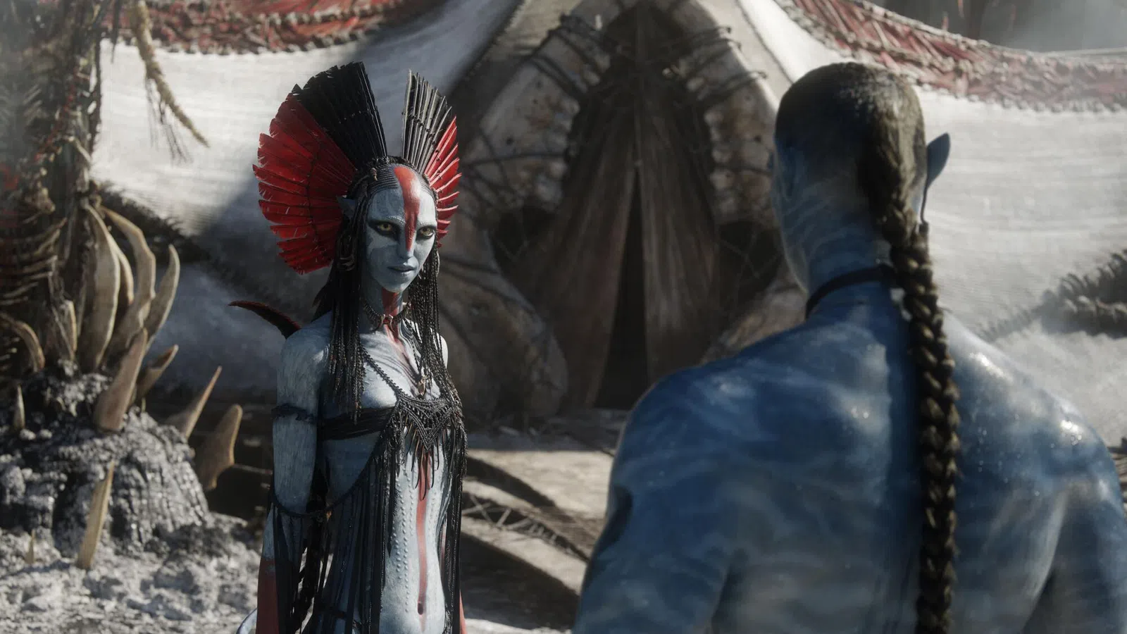 Two blue-skinned Na'vi characters in elaborate costumes face each other in a tribal village setting from Avatar.