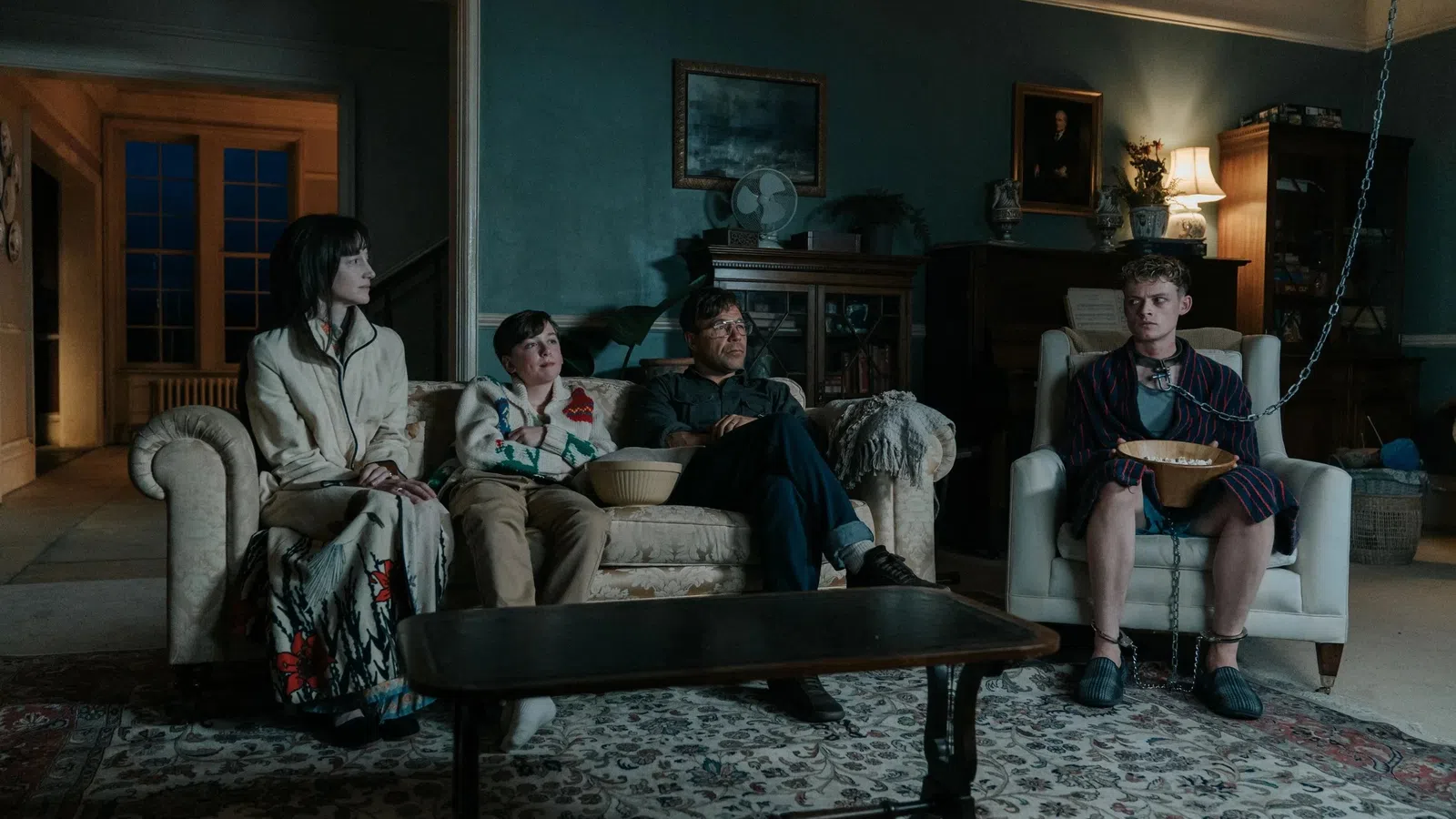 Four people sitting in a dimly lit living room, one chained to a chair holding a bowl.