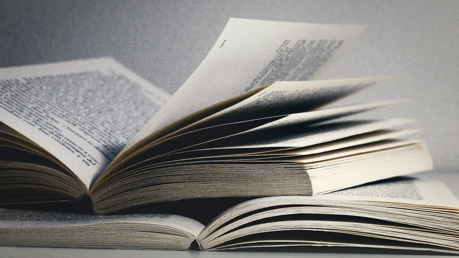 Open books with pages flipping, showing printed text on a neutral background in soft lighting.