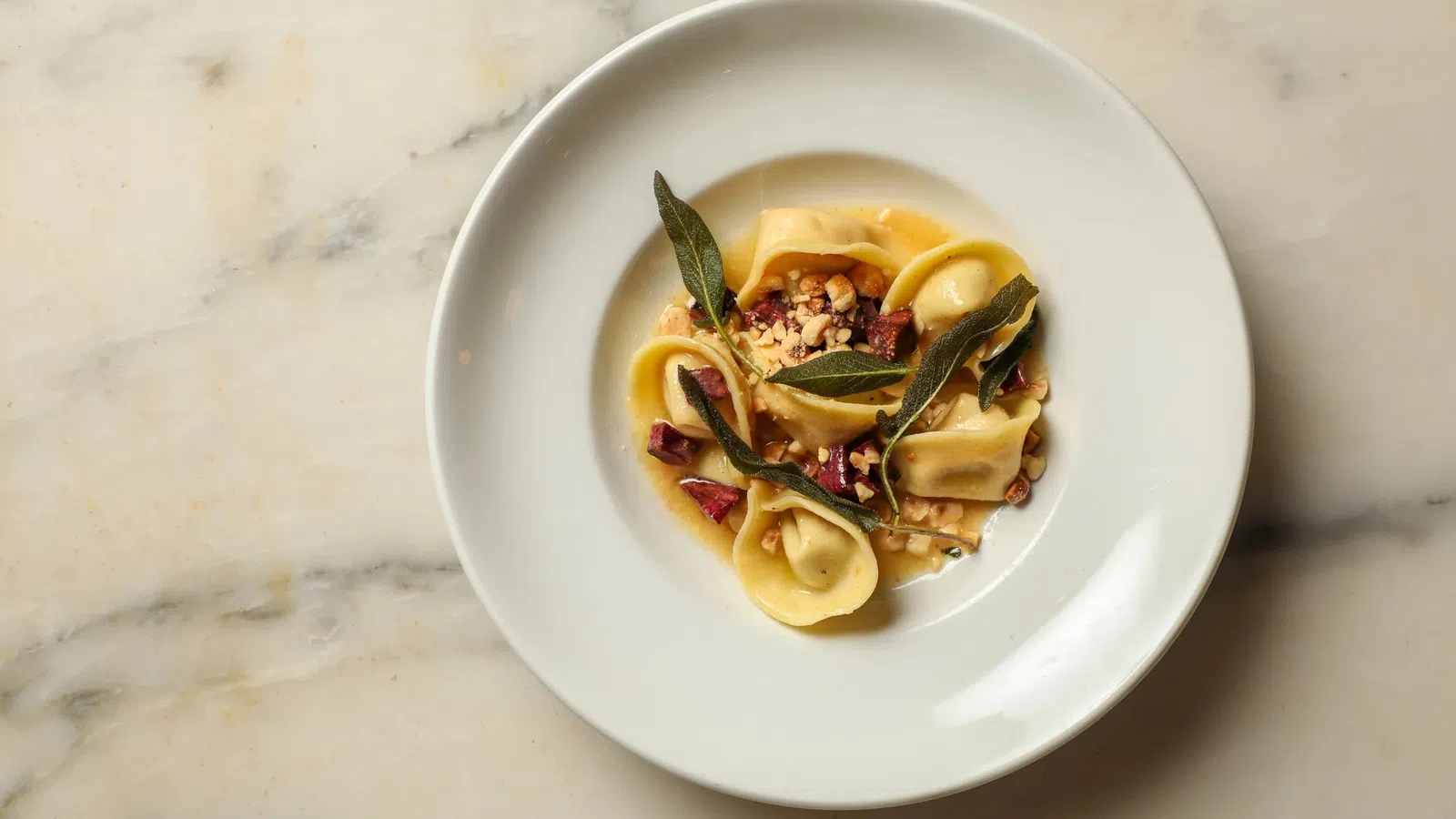 White plate with gourmet tortellini pasta garnished with sage leaves, nuts, and sauce on a marble surface.