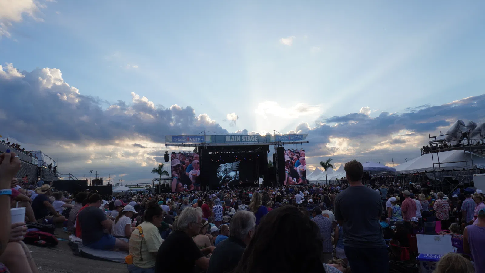 Huge crowd gathered at an outdoor music festival facing the main stage under a partly cloudy sky at sunset.