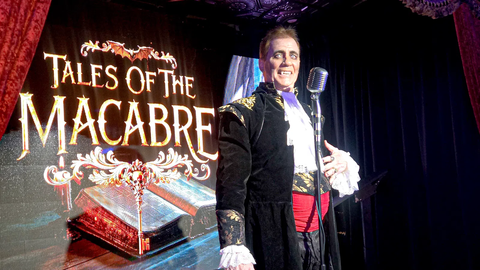 Performer in gothic costume stands next to a microphone with Tales of the Macabre sign behind him on stage.