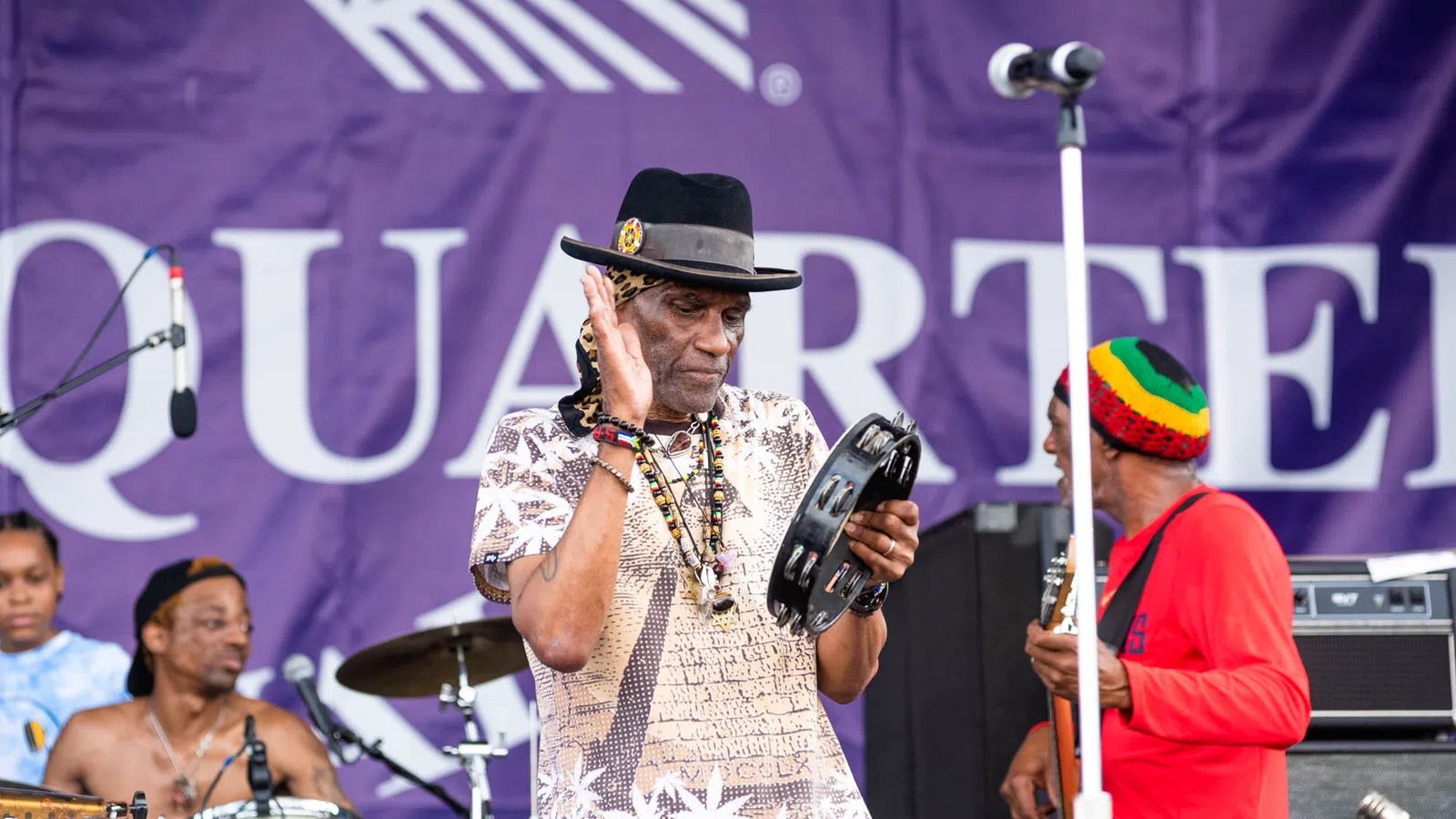 Musicians performing on stage with tambourine and drums, vibrant outfits, and purple backdrop.