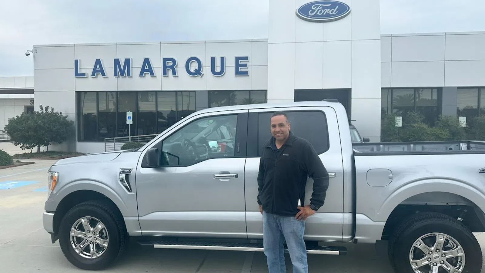 Man standing beside a silver Ford pickup truck in front of Lamarque Ford dealership on a cloudy day