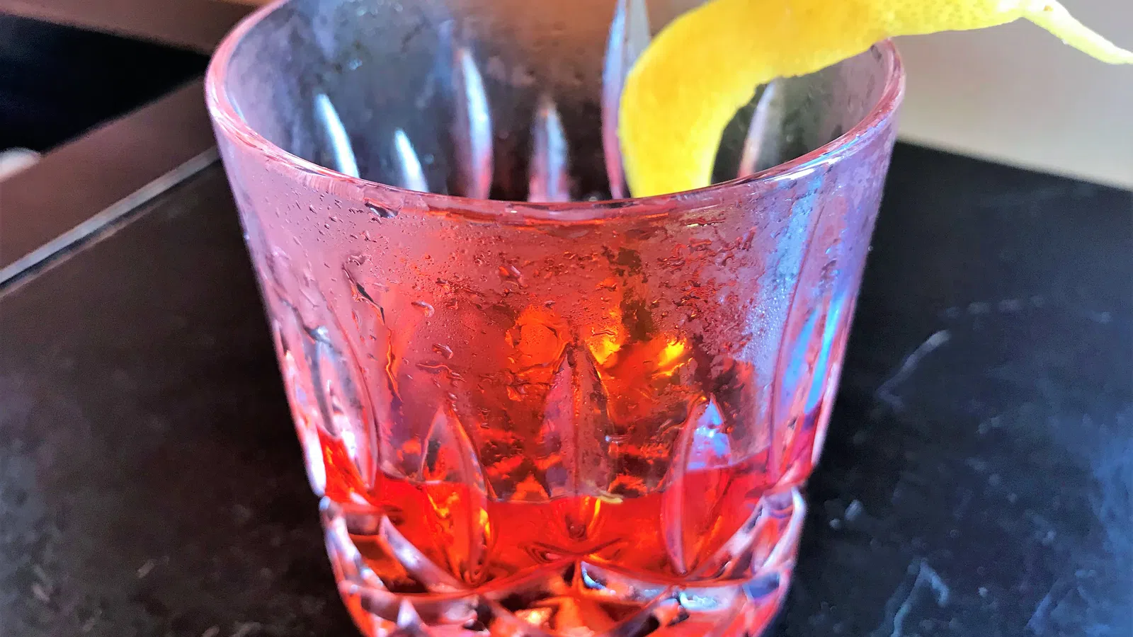Close-up of a classic red cocktail in a textured glass with a lemon twist garnish on dark surface.