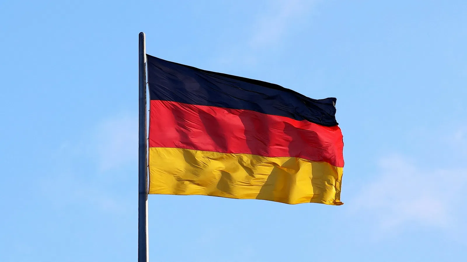 German flag with black, red, and yellow stripes waving on a flagpole against a clear blue sky.