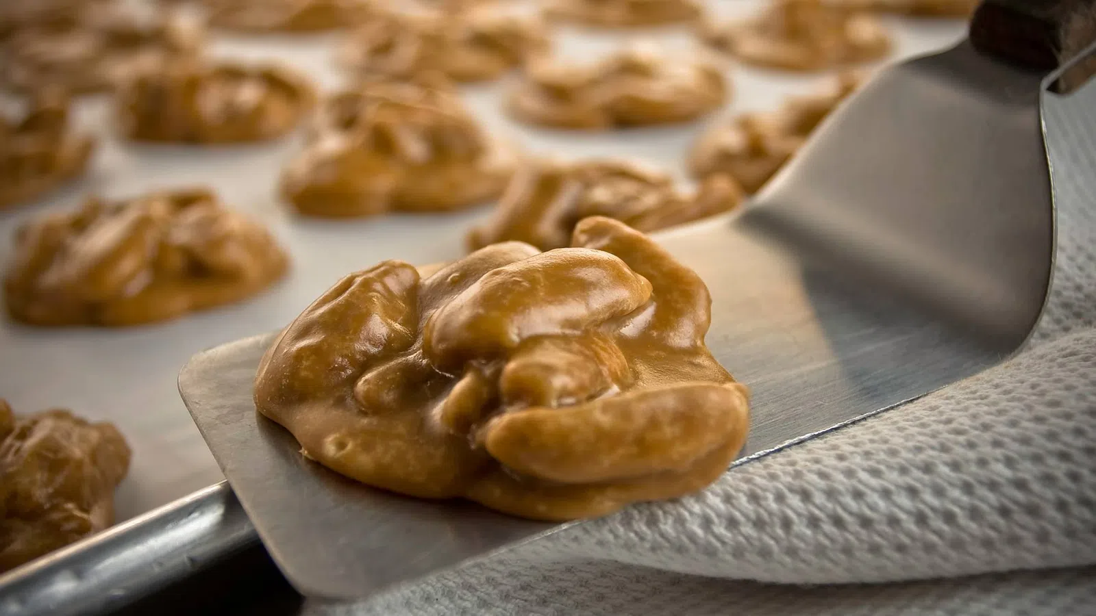 Close-up of freshly made praline candy clusters on a baking sheet being lifted with a metal spatula.