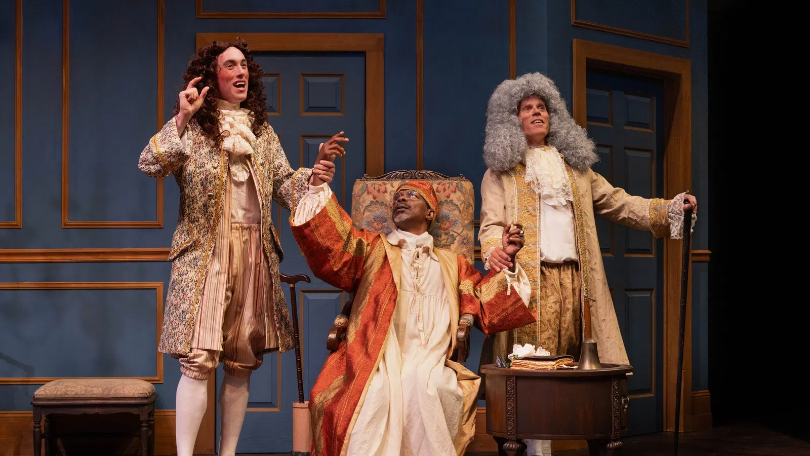 Three actors in 18th-century costumes performing on stage with blue paneled walls and period furniture.